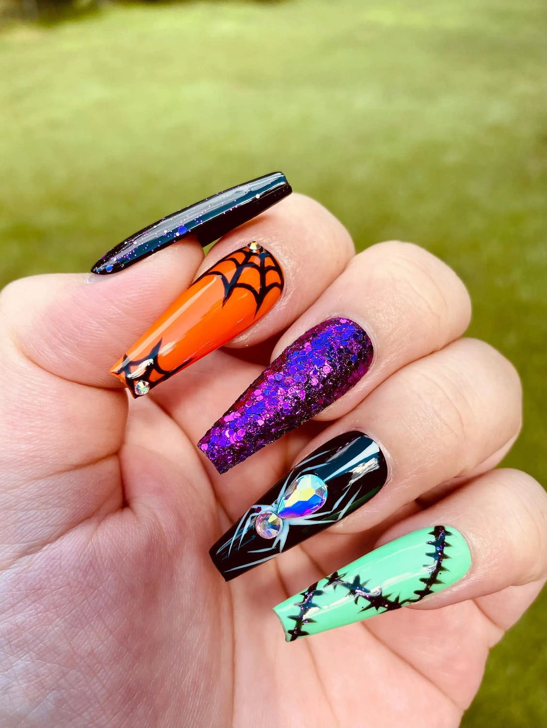 Hand with long press-on nails featuring Halloween designs, glitter, spider web, and rhinestones.