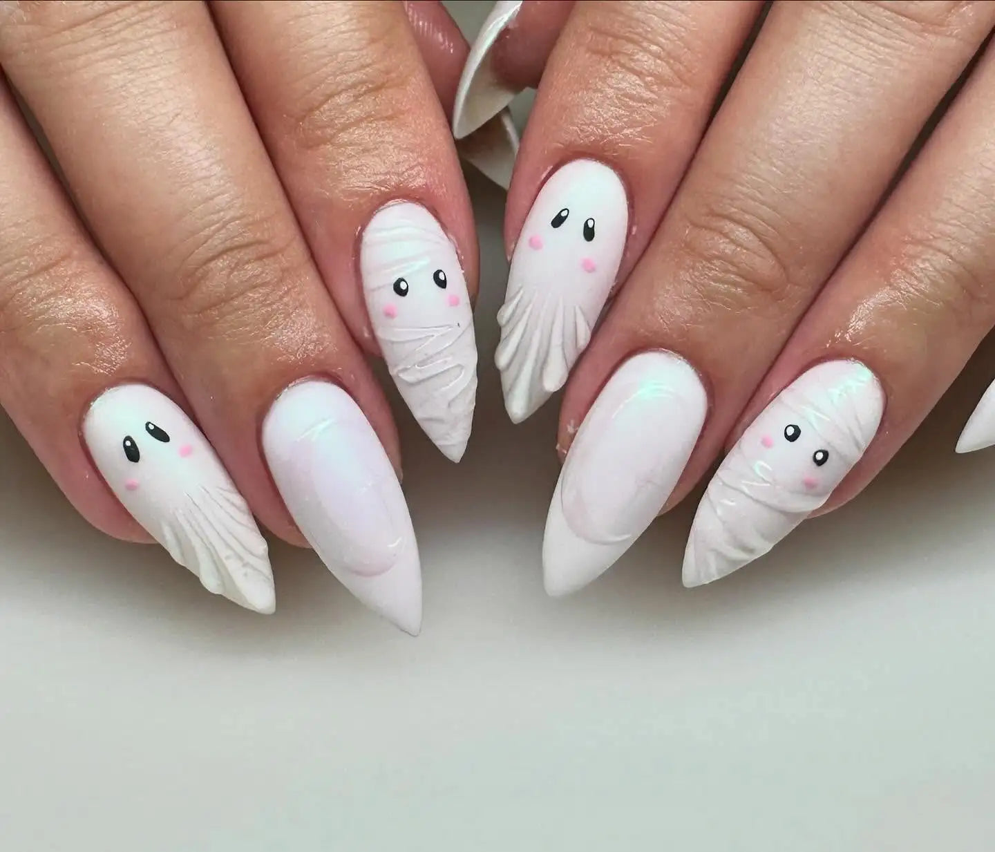 Hand with long press-on nails featuring cute ghost and mummy Halloween nail art in white.