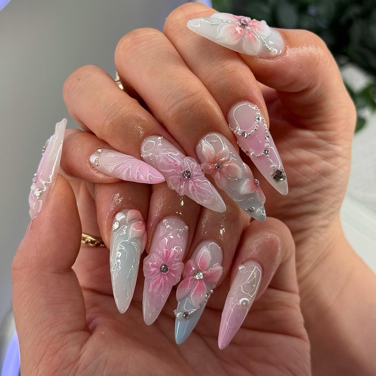 Hand with long, stiletto press-on nails featuring 3D pink floral designs and rhinestone accents.