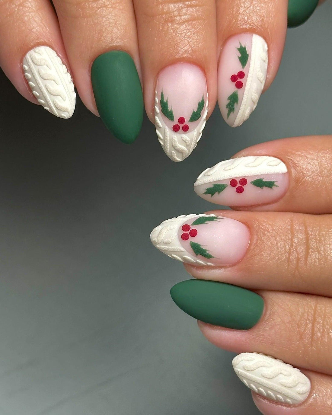 Hand with almond-shaped press-on nails featuring matte green, cable-knit texture, and holly berry holiday nail art.