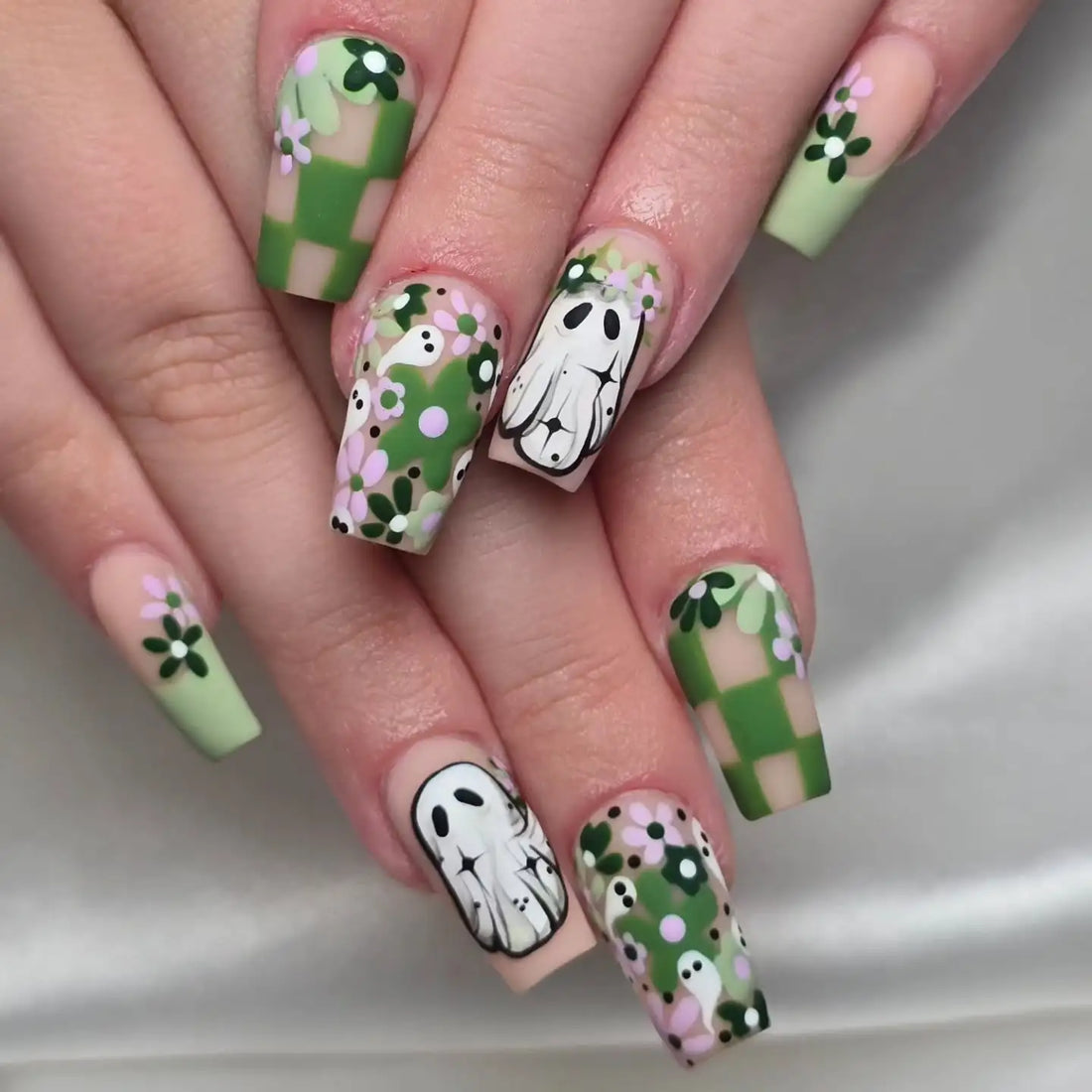 Hand with custom press-on nails featuring green checkerboard, ghost art, and floral designs.