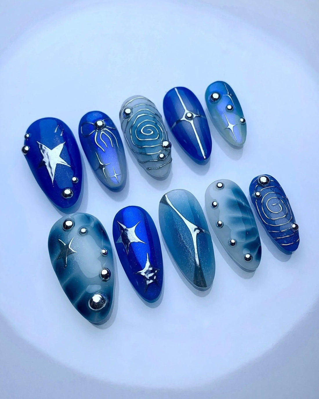 Blue and silver press-on nails with star and swirl designs, metallic accents, and gemstones