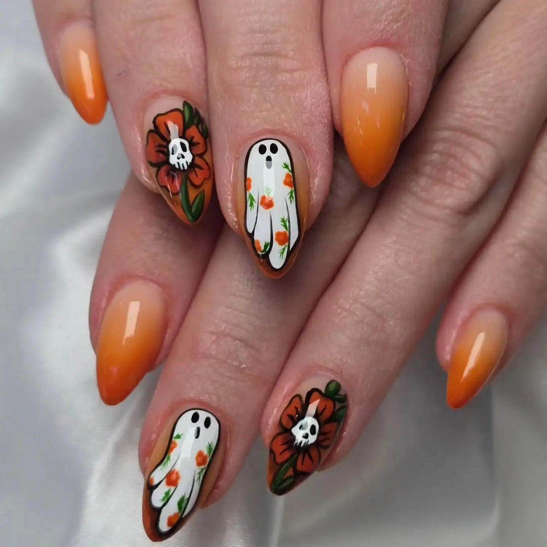 Hand with orange ombre press-on nails featuring floral and ghost nail art designs.