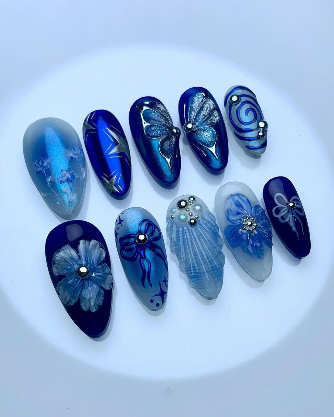 Handmade blue press-on nails with floral, butterfly, and jeweled designs on display.