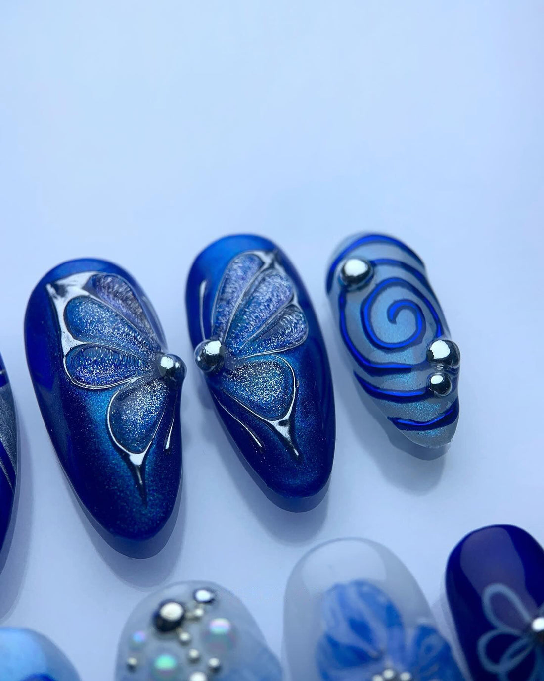Handmade blue press-on nails with floral, butterfly, and jeweled designs on display.