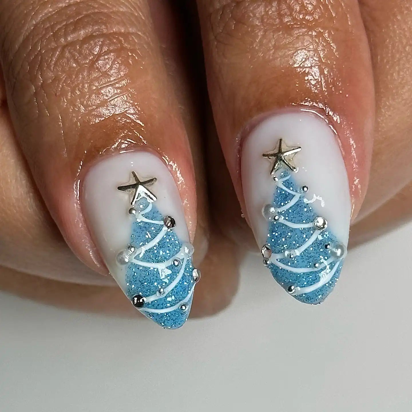 Hand with almond-shaped press-on nails featuring blue glitter Christmas tree nail art with star toppers.