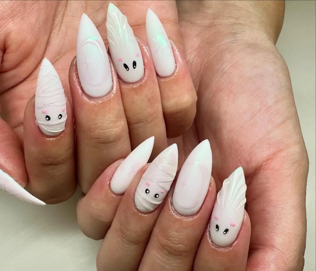 Hand with long press-on nails featuring cute ghost and mummy Halloween nail art in white.
