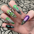 Hand with Halloween press-on nails featuring green glitter, black and white stripes, spider web charm, and purple nails with glitter accents.