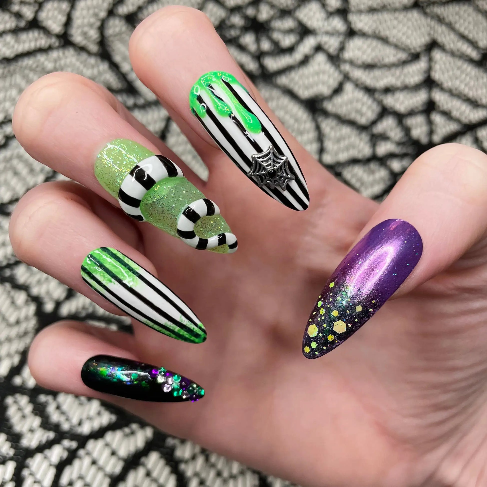 Hand with Halloween press-on nails featuring green glitter, black and white stripes, spider web charm, and purple nails with glitter accents.