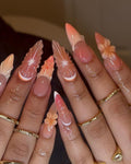Long press-on nails with peach and nude butterfly and flower designs, worn with gold rings.