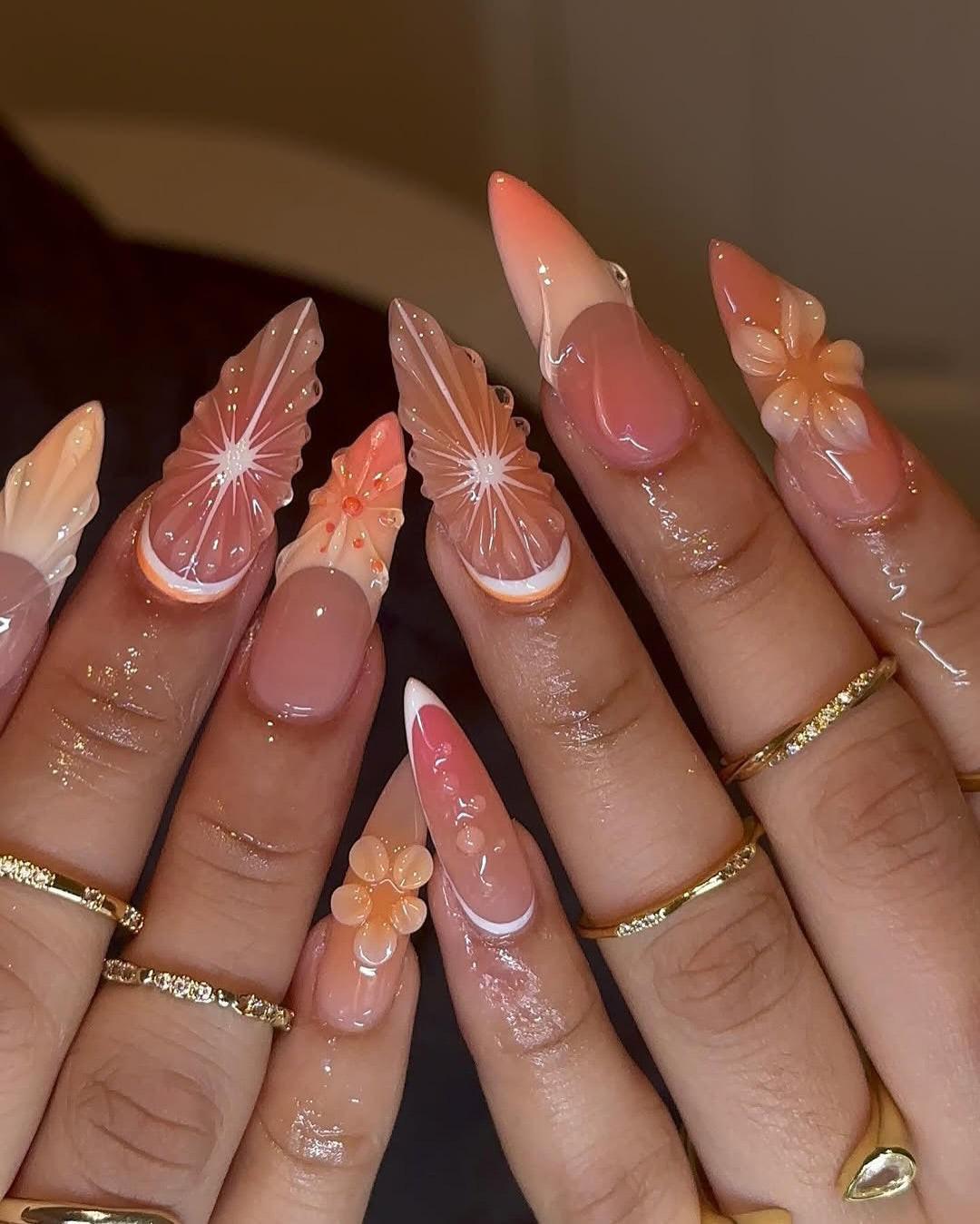 Long press-on nails with peach and nude butterfly and flower designs, worn with gold rings.