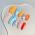 Handmade press-on nails with citrus fruit, blue marble, and floral designs on a neutral background.