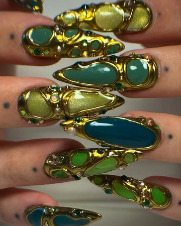 Hand with long press-on nails featuring gold, green, and teal gemstone designs