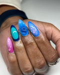 Hand with vibrant handmade press-on nails in blue, teal, and pink, featuring hibiscus and water droplet designs.