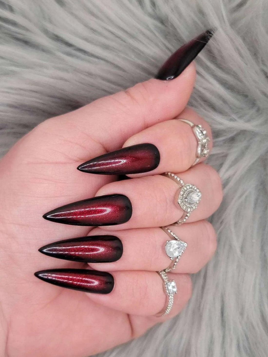 Long stiletto black and red ombre press-on nails with gothic style, worn with silver rings