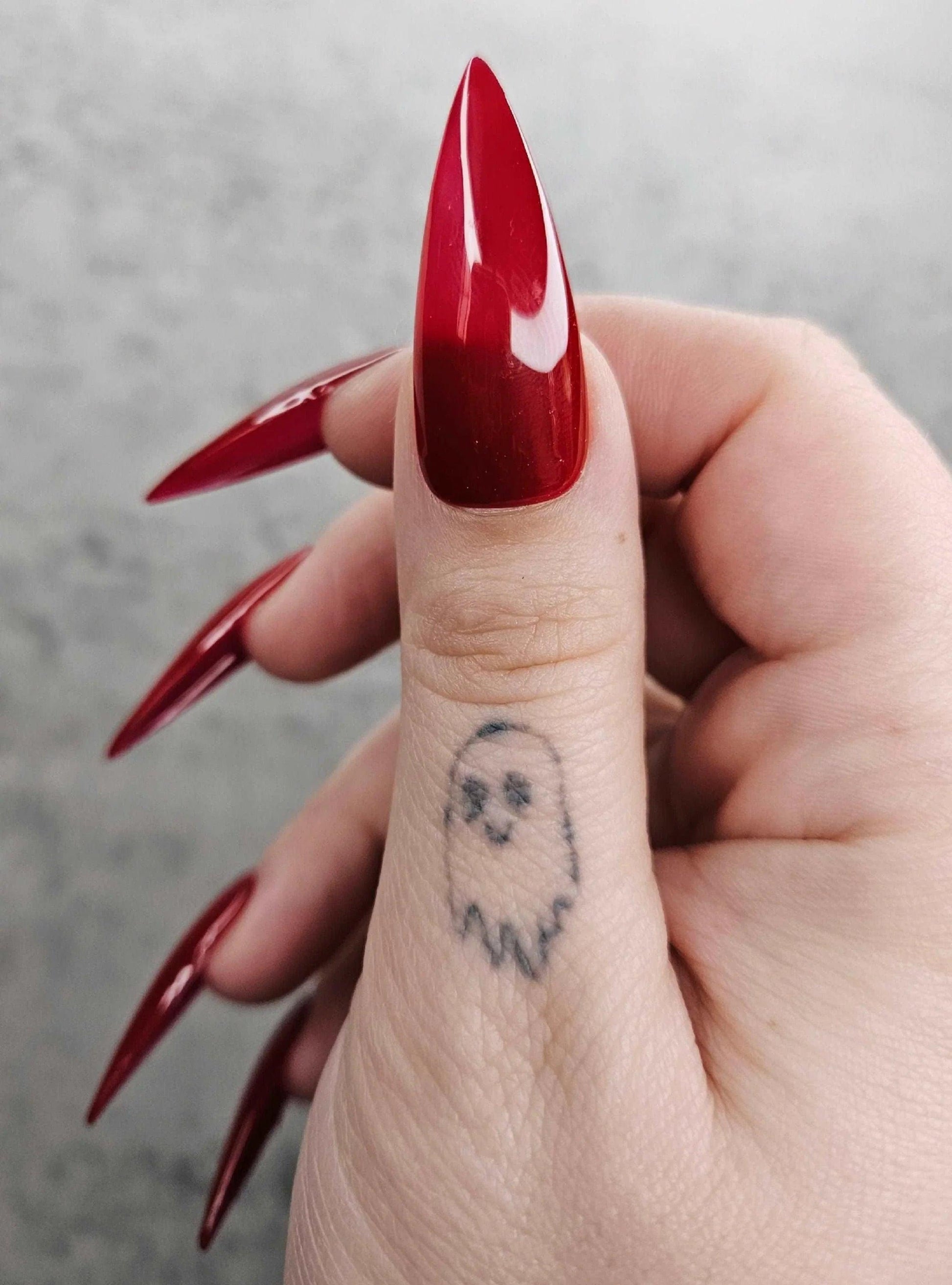 Hand with long, glossy stiletto red press-on nails and a small ghost tattoo on the thumb.