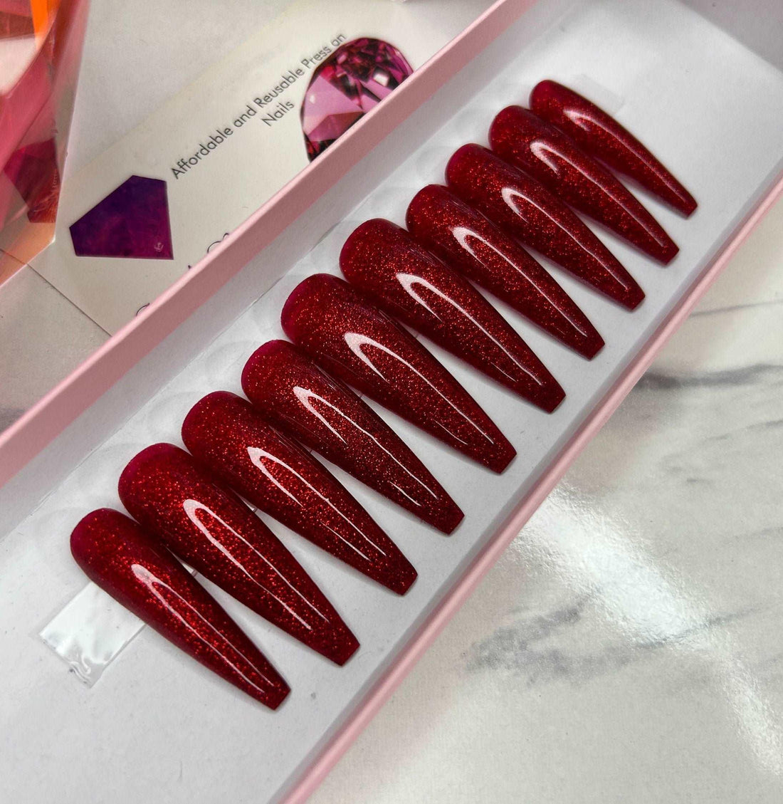 Coffin-shaped glittery red press-on nails in a pink box on a marble surface