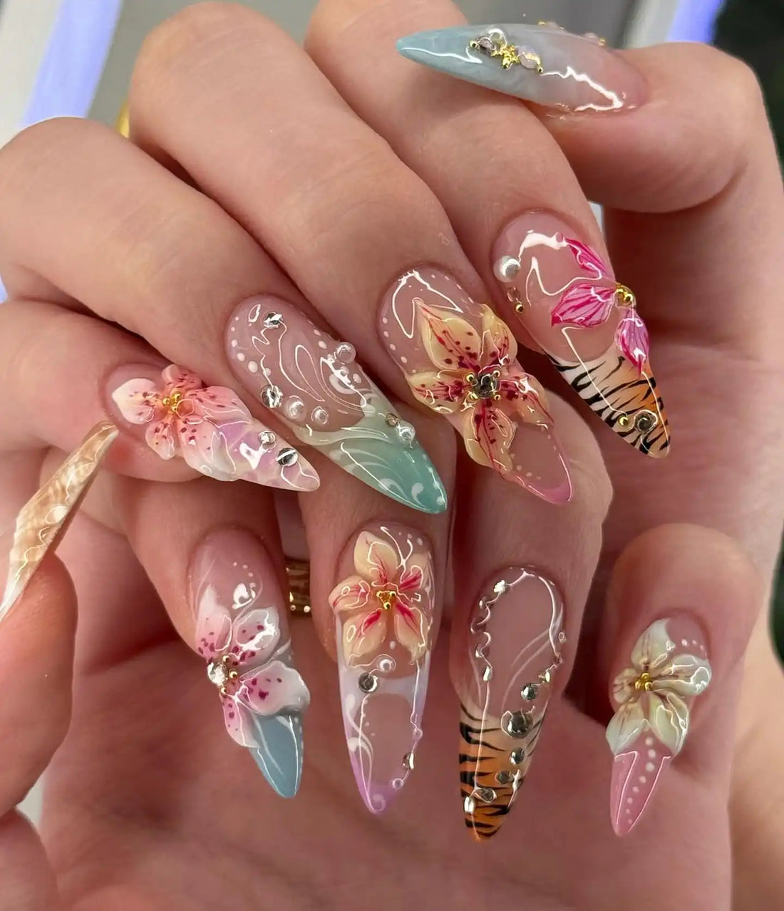Hand holding long stiletto press-on nails with 3D floral designs, ombre colors, tiger print, and rhinestones