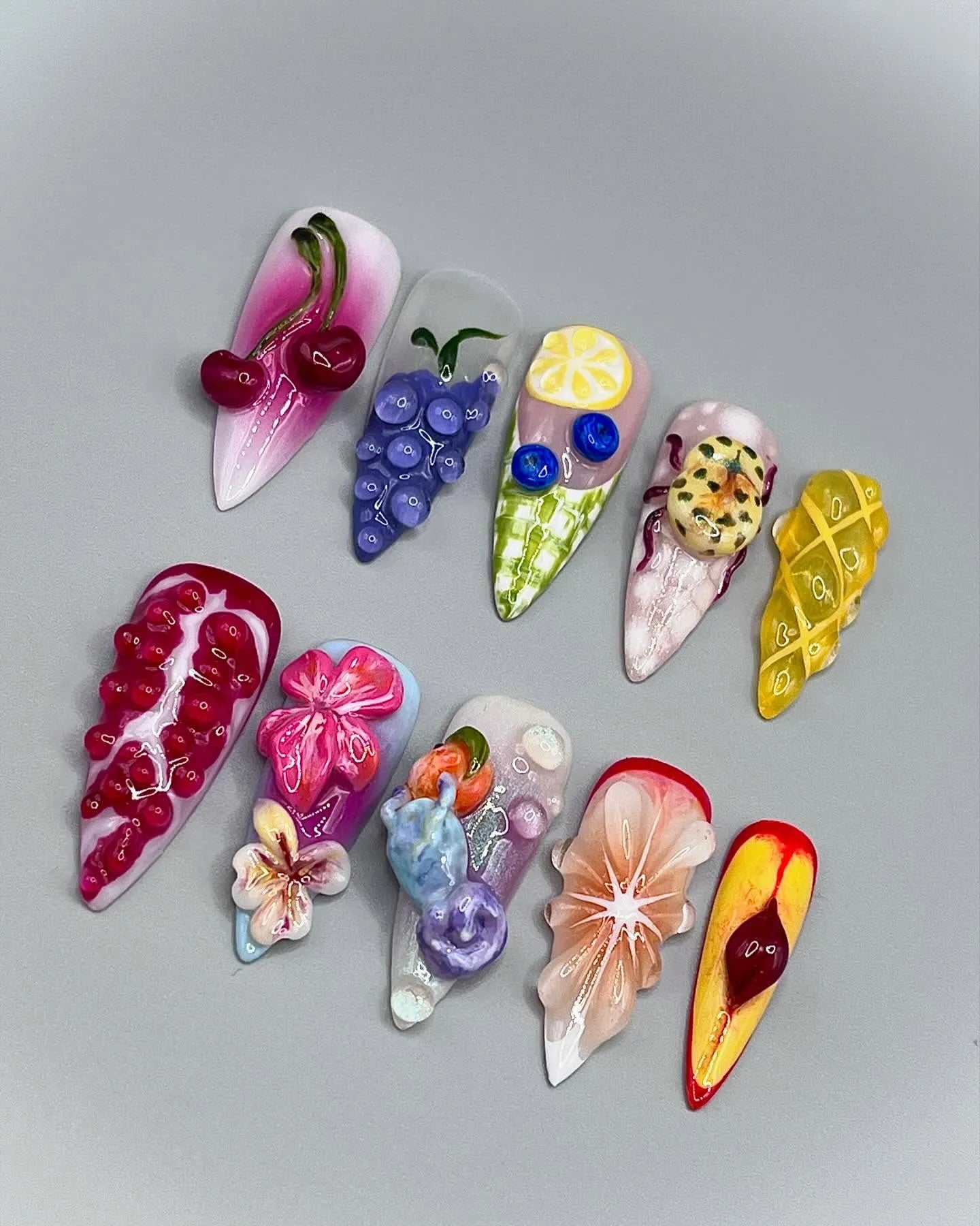 Handmade 3D press-on nails with fruit and floral designs, including cherry, lemon, pomegranate, and flower art, displayed on a gray background.