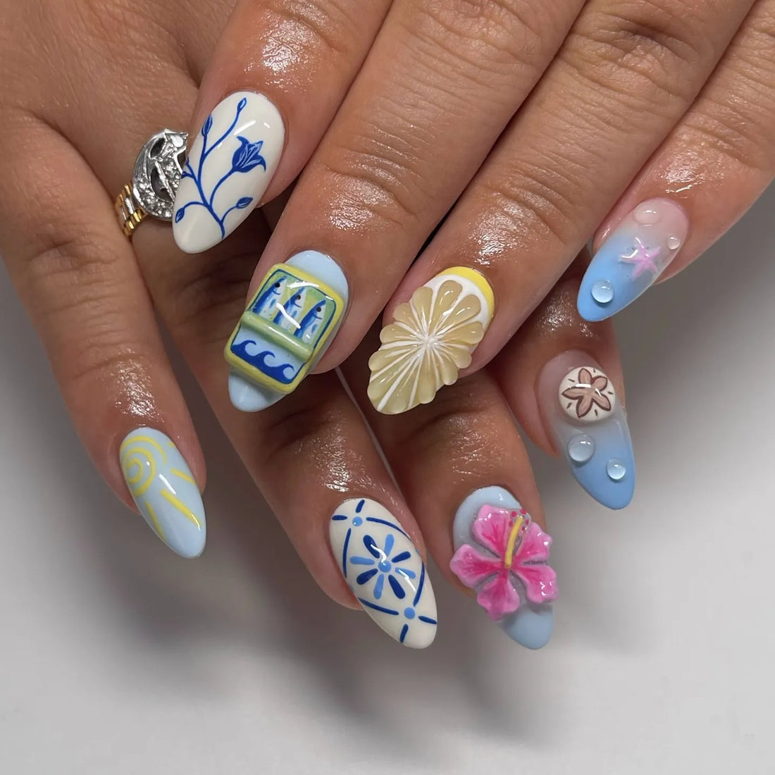 Hand with blue and white 3D floral summer vacation press-on nails featuring lemon, hibiscus, seashell, and ocean designs.