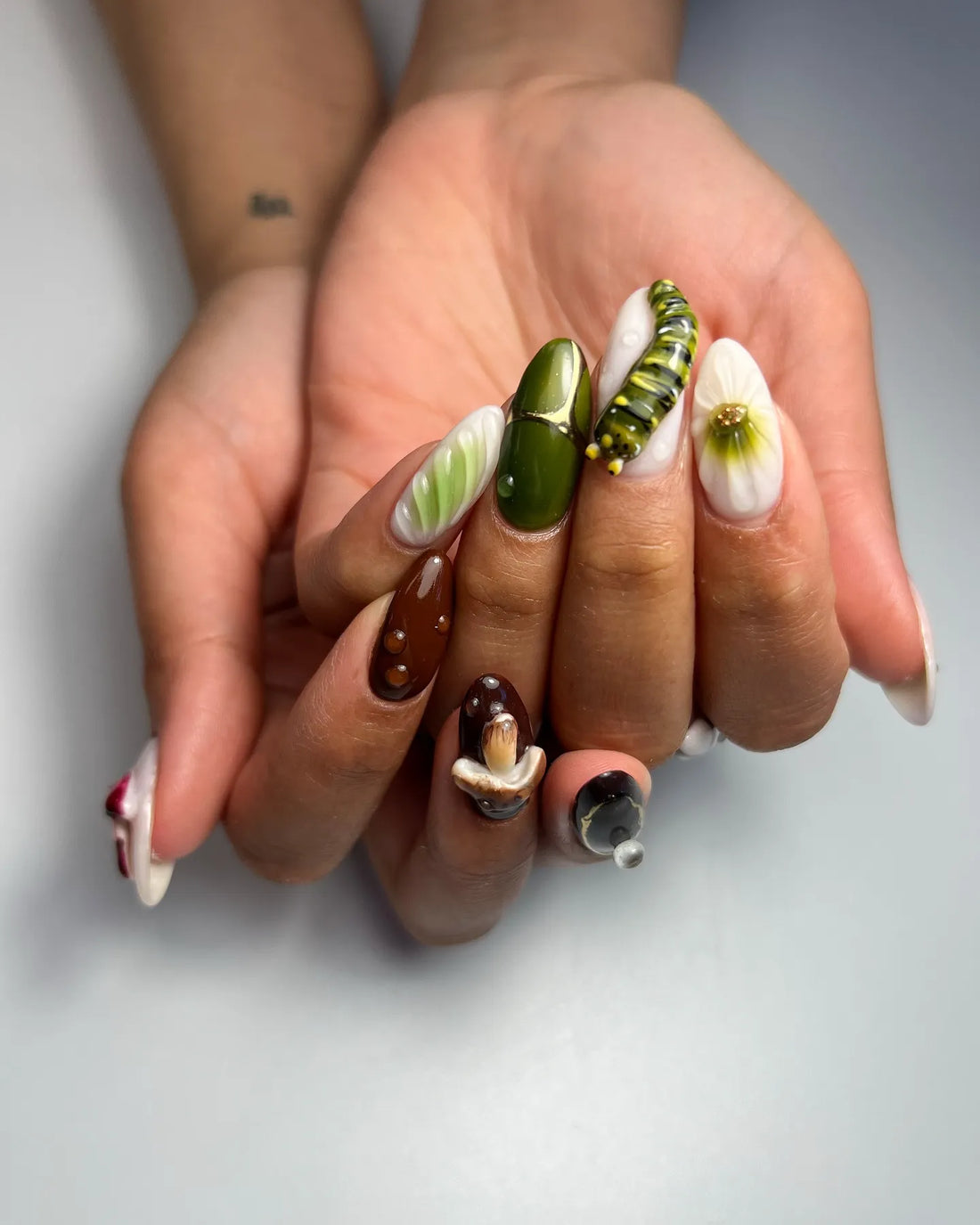 Hand with handmade 3D press-on nails featuring mushroom, caterpillar, and forest designs in green and brown.