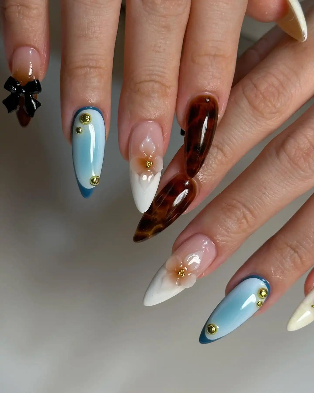 Hand with long stiletto press-on nails featuring brown, blue, and white designs, 3D flowers, gold studs, and a black bow.
