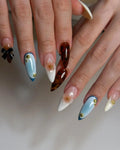 Hand with stiletto-shaped handmade press-on nails in blue, white, and tortoiseshell with gold accents, 3D flowers, and a black bow design.