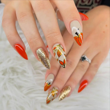 Hand with autumn press-on nails featuring pumpkin, leaf, orange, and gold glitter designs
