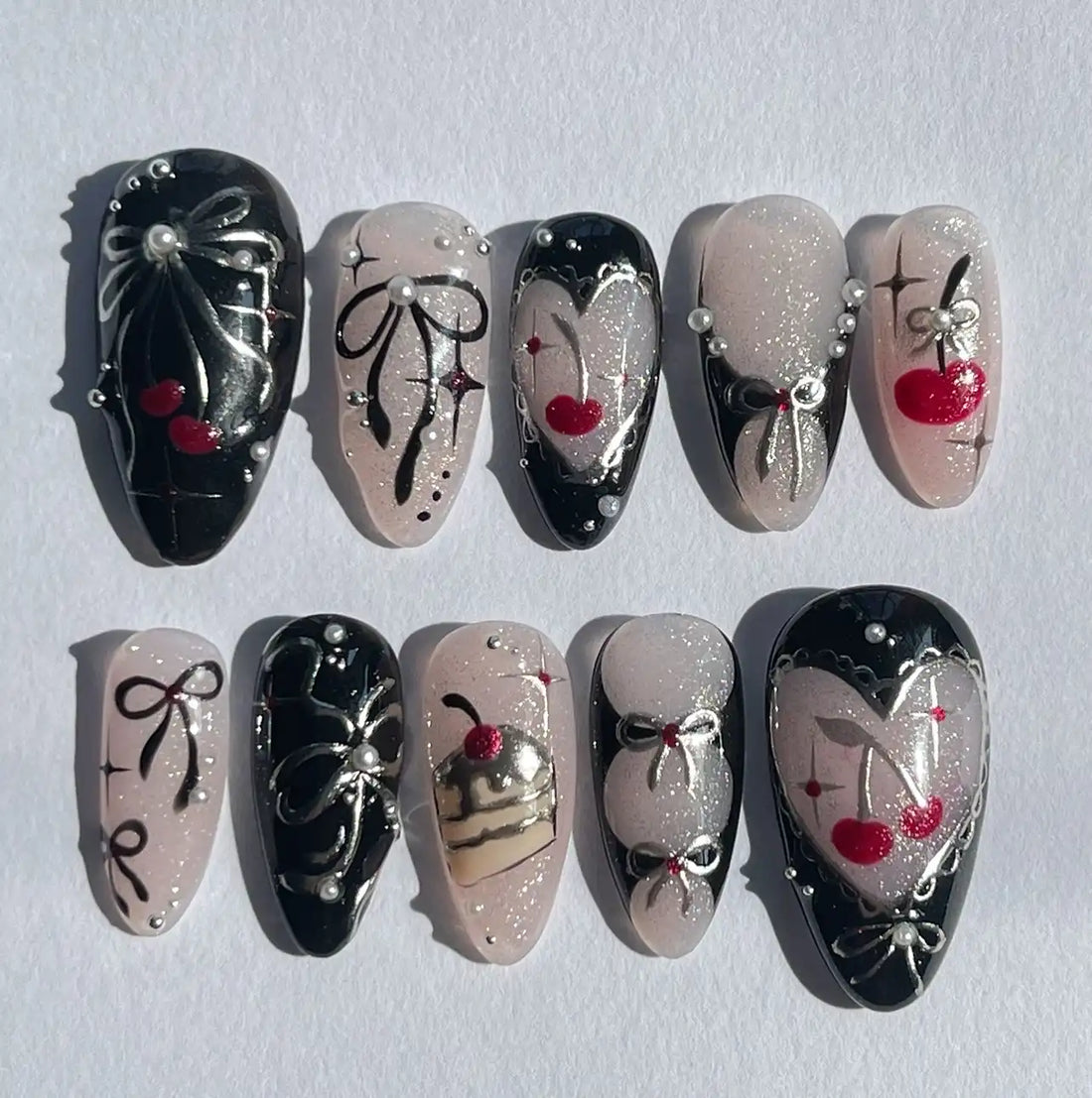 Handmade press-on nails with black and pink designs, cherries, bows, pearls, and glitter accents.