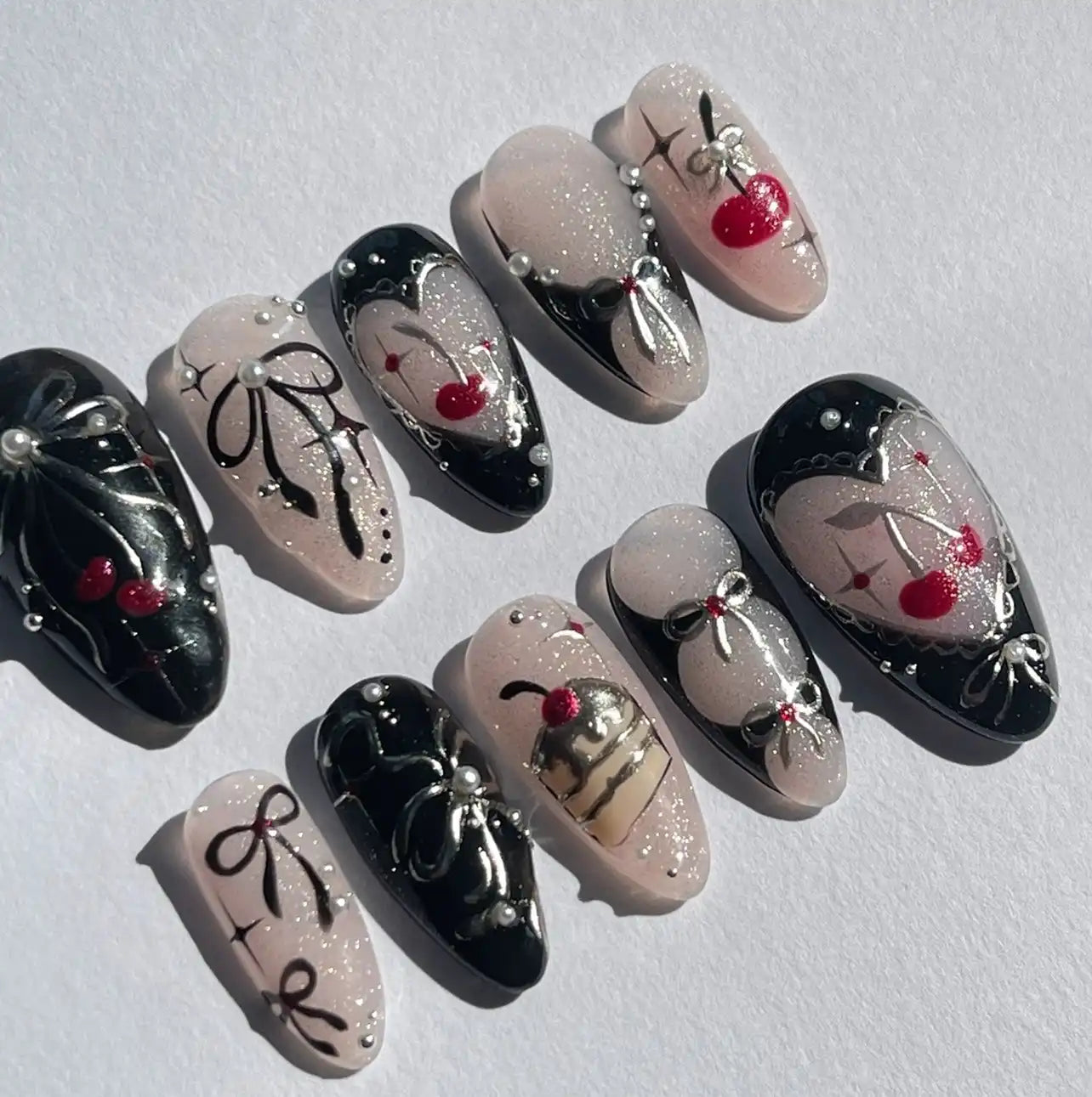Handmade press-on nails with black and pink designs, cherry art, bows, pearls, and glitter accents.