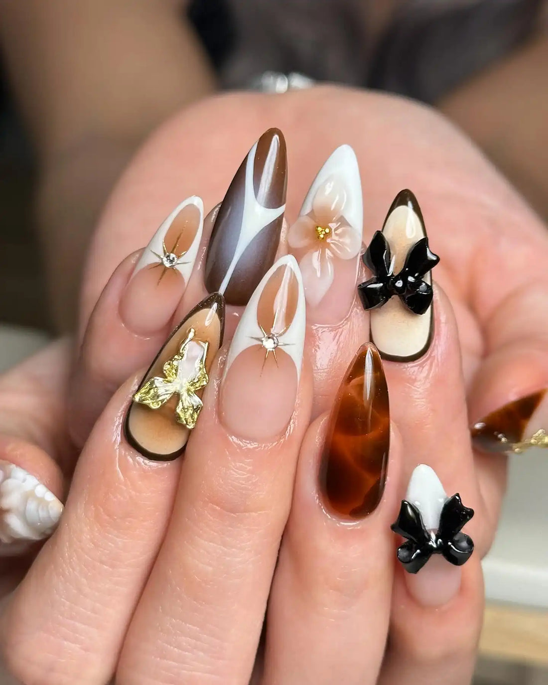 Handmade press-on nails with brown marble, gold accents, black 3D bows, and floral designs.