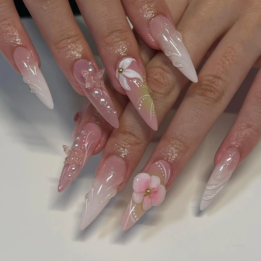 Long stiletto press-on nails with 3D flowers, butterfly accents, pearls, and pastel designs.