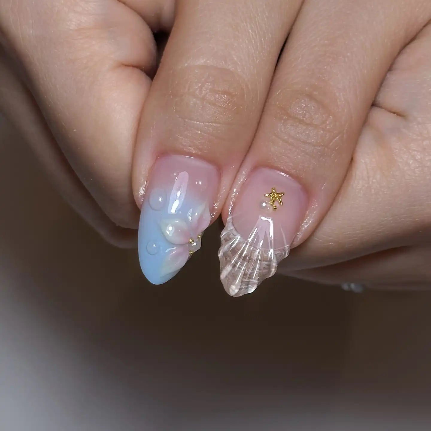 Hand holding handmade press-on nails with seashell designs, pink and blue gradient, pearl accents