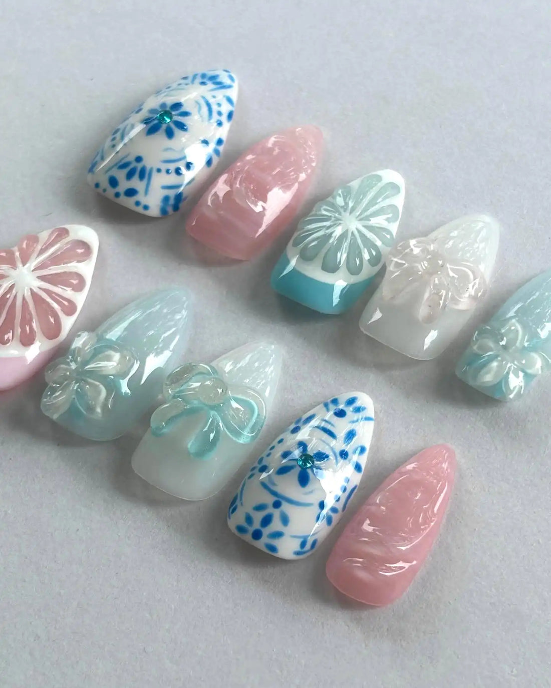 Handmade press-on nails with blue floral, pastel bow, and citrus designs on a gray background.