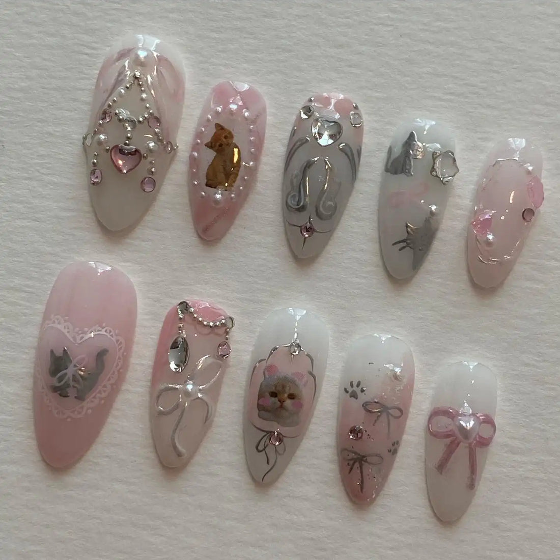 Handmade pink and white press-on nails with cat designs, pearls, bows, rhinestones, and lace accents.