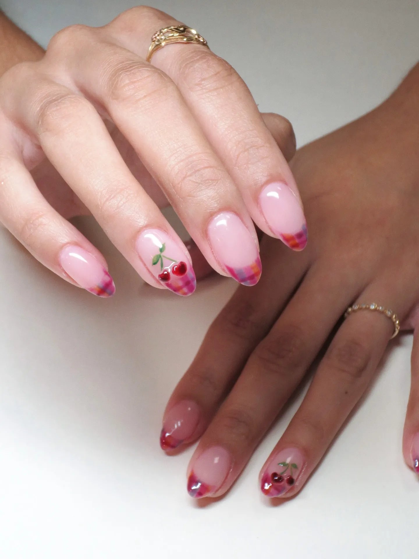 Hand with almond-shaped press-on nails featuring pink plaid French tips and cherry fruit nail art.