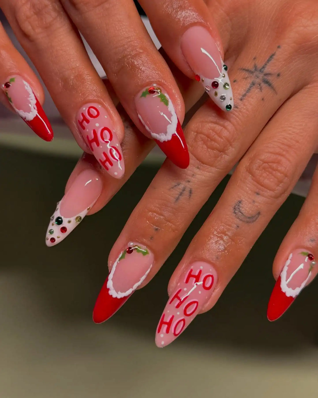 Hand with Christmas press-on nails featuring red tips, holly, and HO HO HO festive designs.