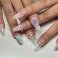 Hand with long handmade press-on nails featuring 3D floral and butterfly designs in pastel colors.
