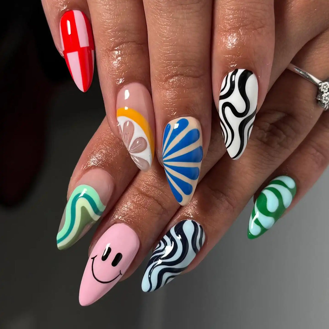 Hand with colorful handmade press-on nails featuring swirls, patterns, and a smiley face design.