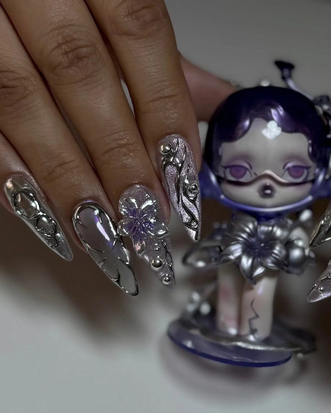 Hand with stiletto press-on nails in chrome and purple, featuring 3D metallic floral designs.