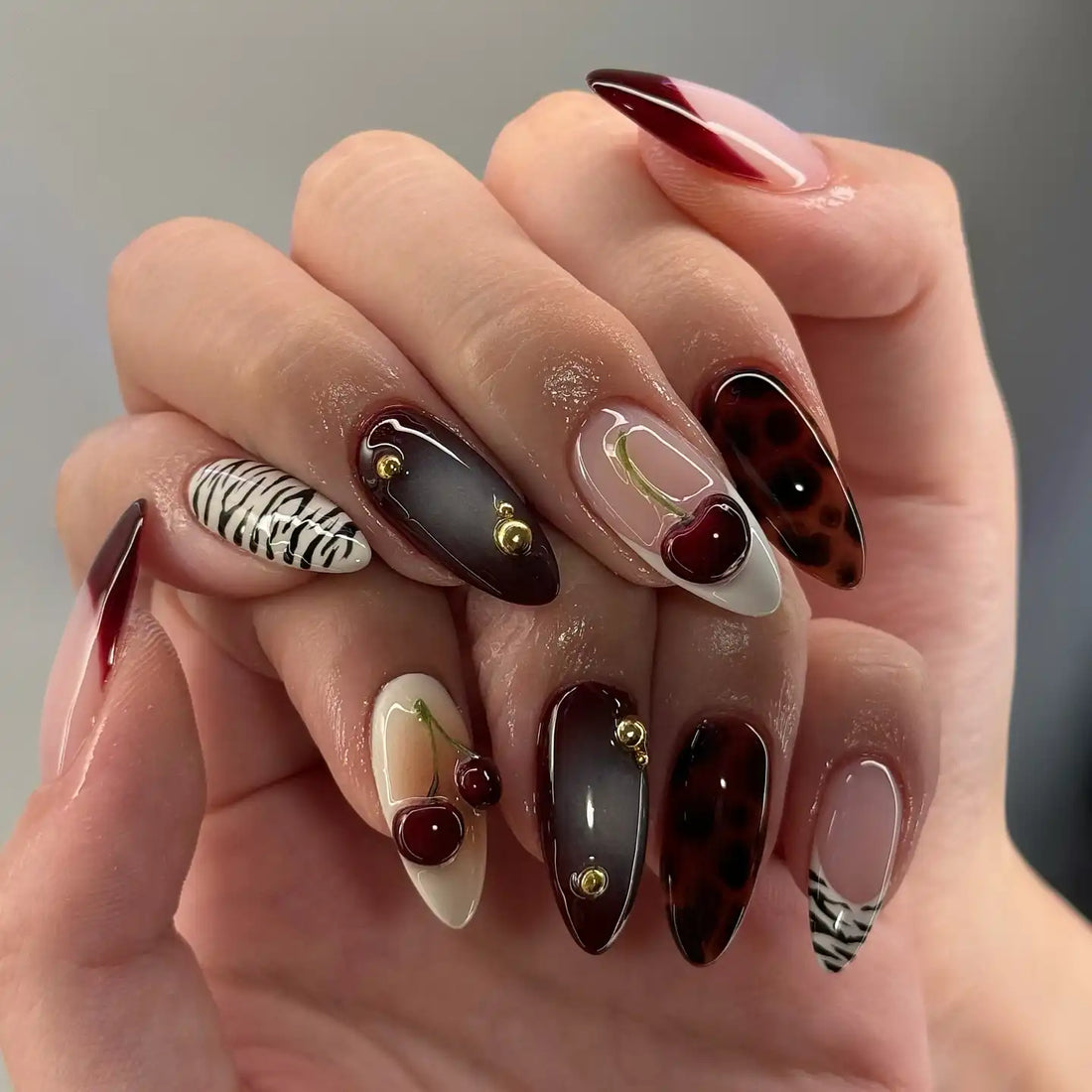 Hand holding handmade deep red 3D cherry and gold pearl press-on nails with zebra accents.