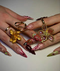 Hand with long press-on nails in deep red and pastel designs, decorated with 3D floral art and gemstones.