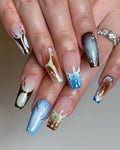 Hand with handmade ombre press-on nails in blue, brown, and silver designs with rhinestones.
