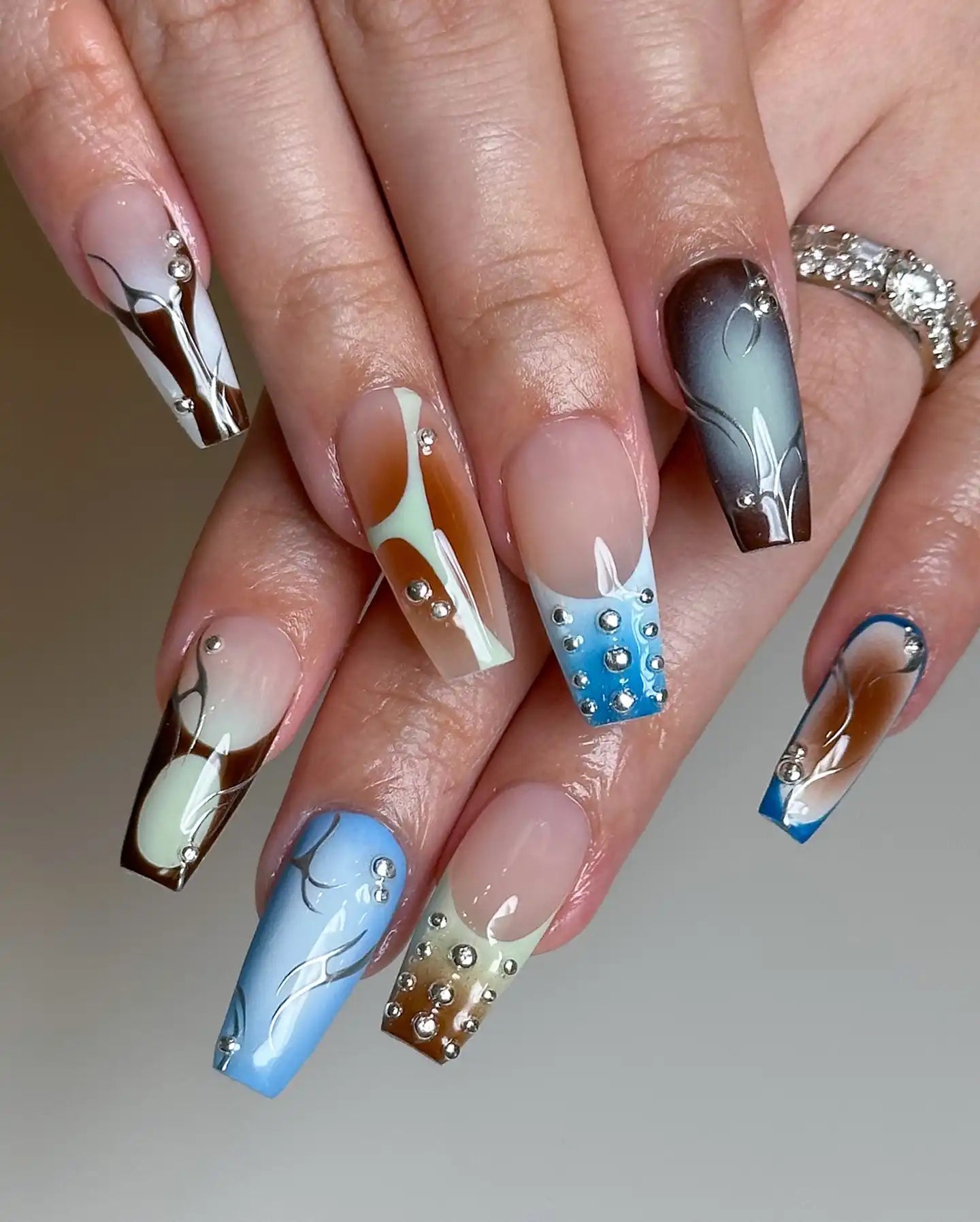 Hand with handmade ombre press-on nails in blue, brown, and silver designs with rhinestones.