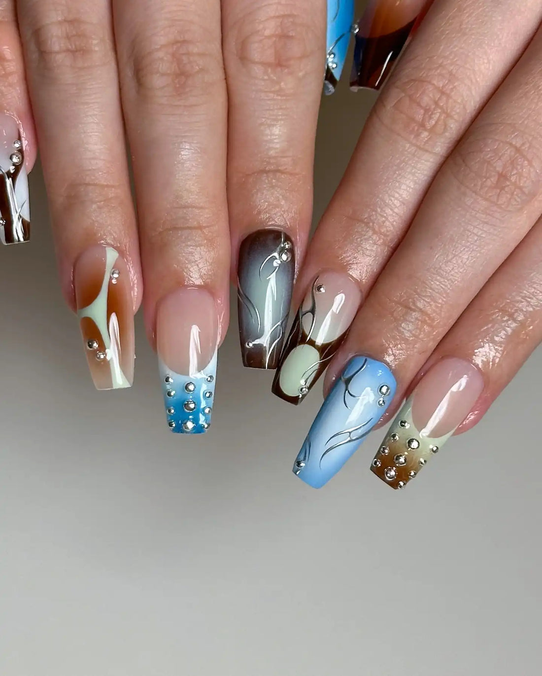 Hand with handmade ombre press-on nails in blue, brown, and silver designs with rhinestones.