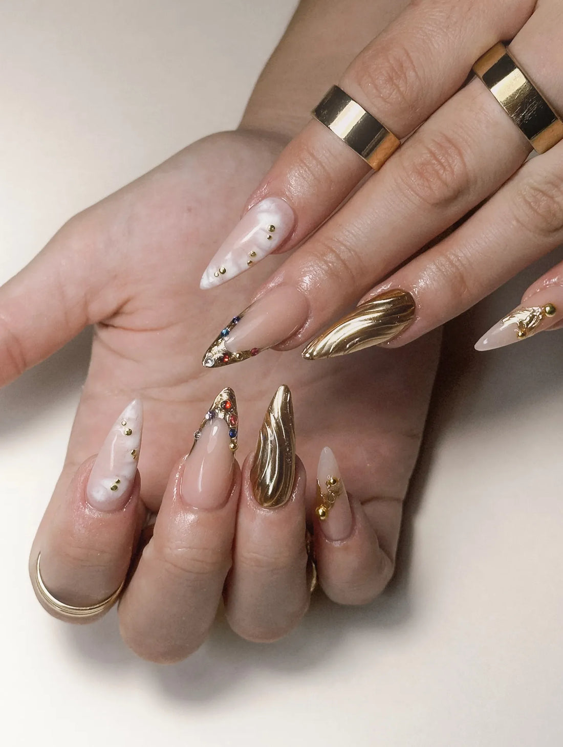 Hand with long stiletto press-on nails featuring gold chrome, marble white, and multicolor gemstones.