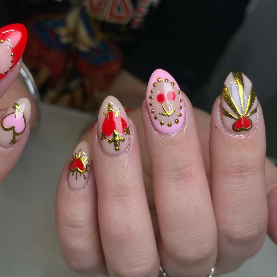 Handmade press-on nails with red hearts, gold dagger, cherry, and gothic designs on nude base.