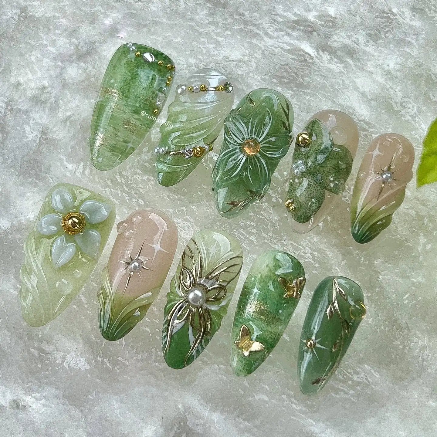 Handmade green floral press-on nails with pearl and butterfly accents on textured background.