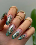 Hand with long handmade green floral stained glass press-on nails, gold rings, nature-inspired design.