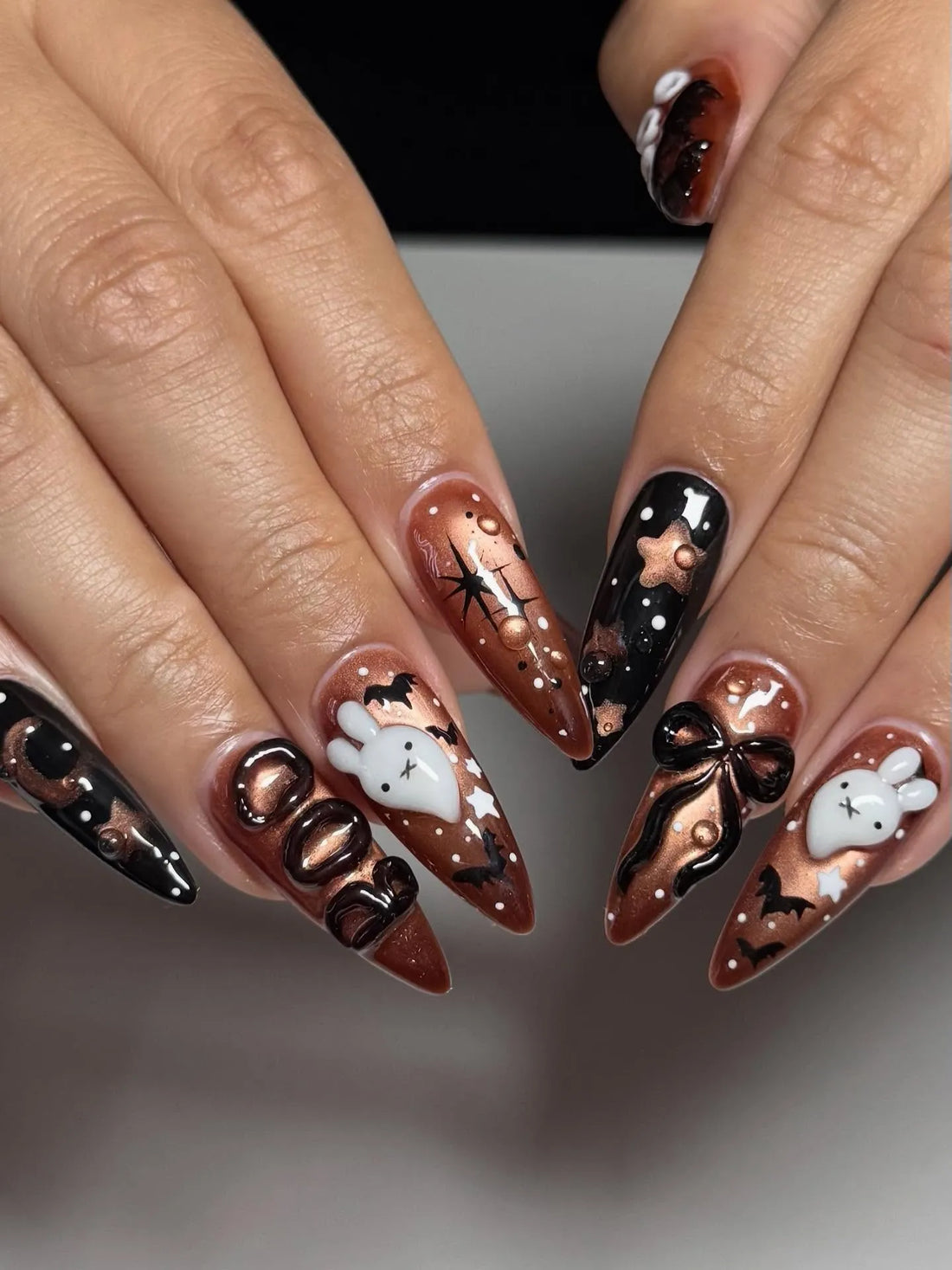 Hand with long stiletto press-on nails in black and bronze, decorated with 3D Halloween ghost, bow, bats, and spiderweb designs.